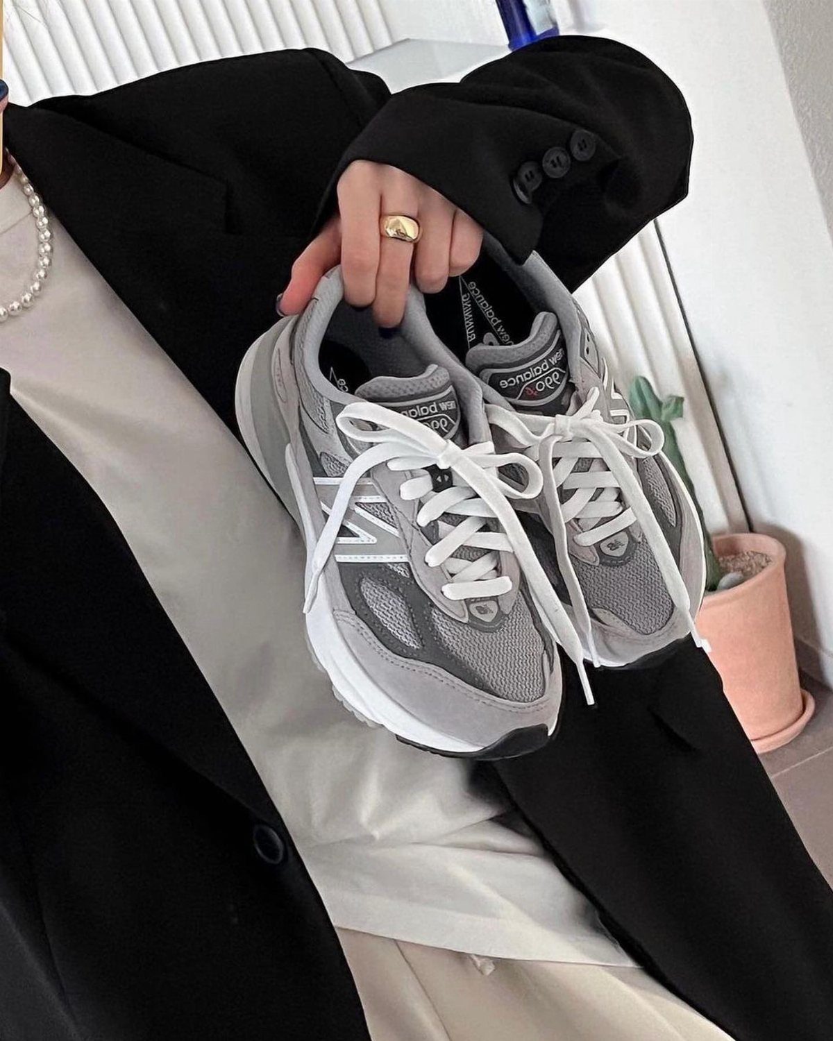 A person in a black blazer holding a pair of grey New Balance athletic sneakers with white laces.