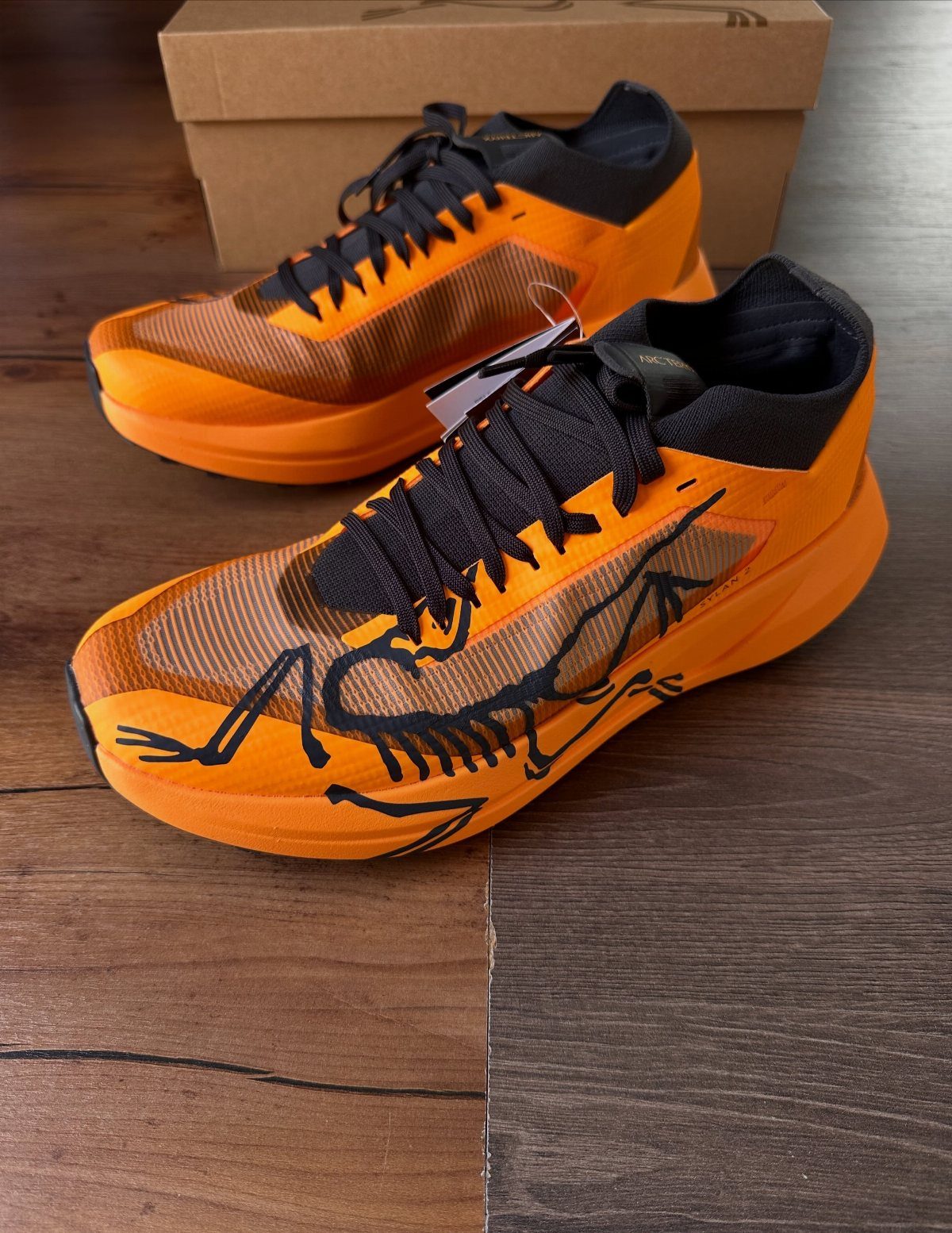 Orange athletic shoes with black skeletal graphics sitting on a wooden floor near a box.