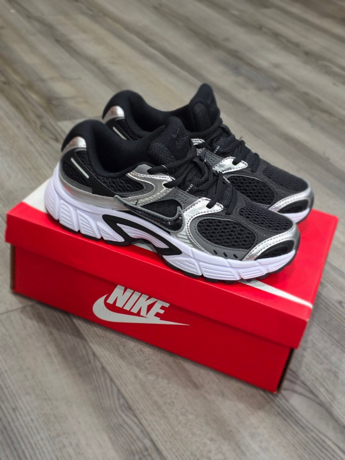 Black and silver sneakers resting on a red Nike shoebox on a wooden floor.