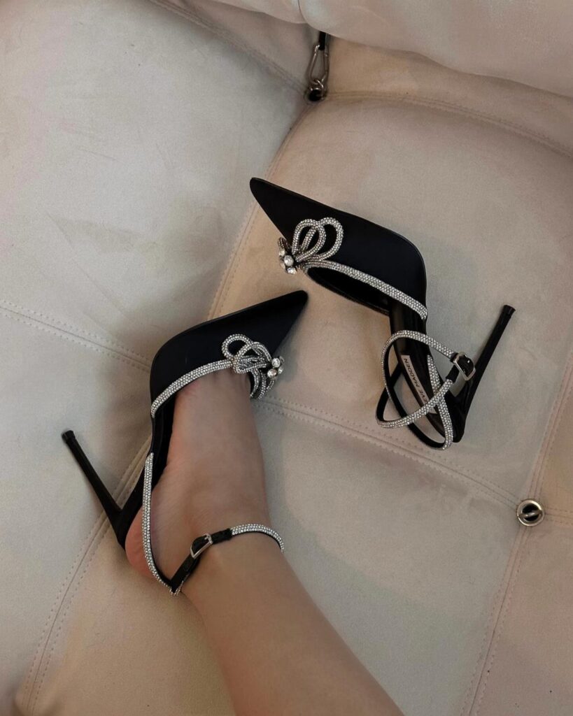 Black pointed-toe high heels with silver rhinestone straps worn on a person's foot.