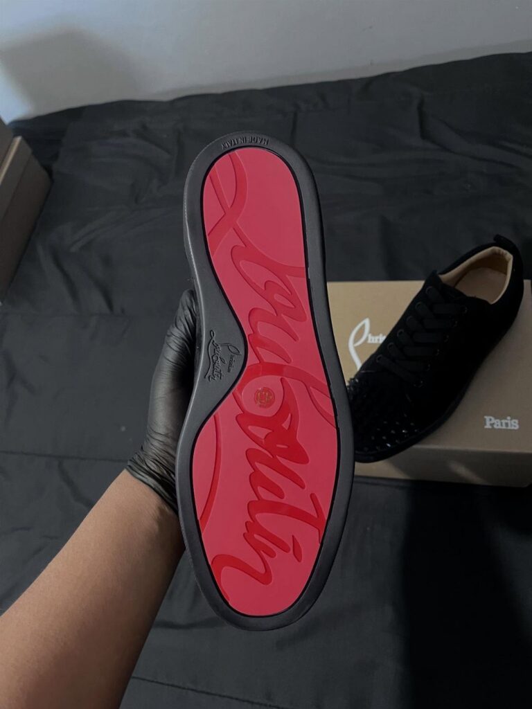 A gloved hand holds a black sneaker displaying a bright red designer sole.