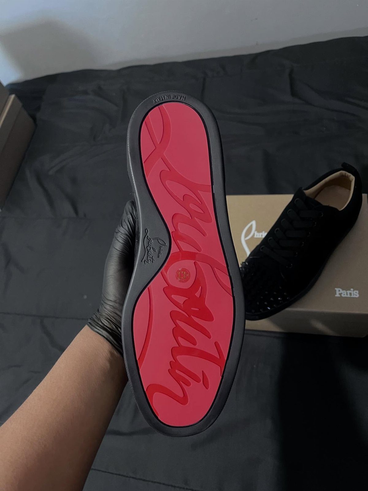 A gloved hand holds a black sneaker displaying a bright red designer sole.