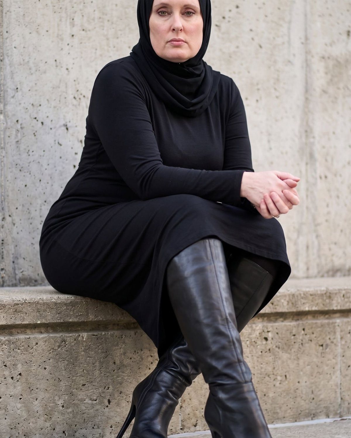 A woman in a black dress and hijab wearing tall black leather boots.