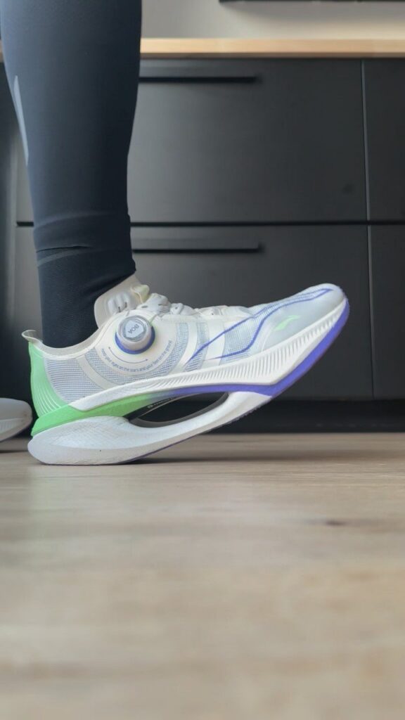A white and blue racing running shoe with a thick green and purple midsole on a wooden floor.