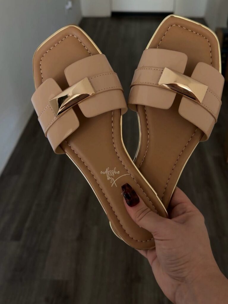 Tan flat open-toe slide sandals with wide straps and gold metallic buckles held by a hand.