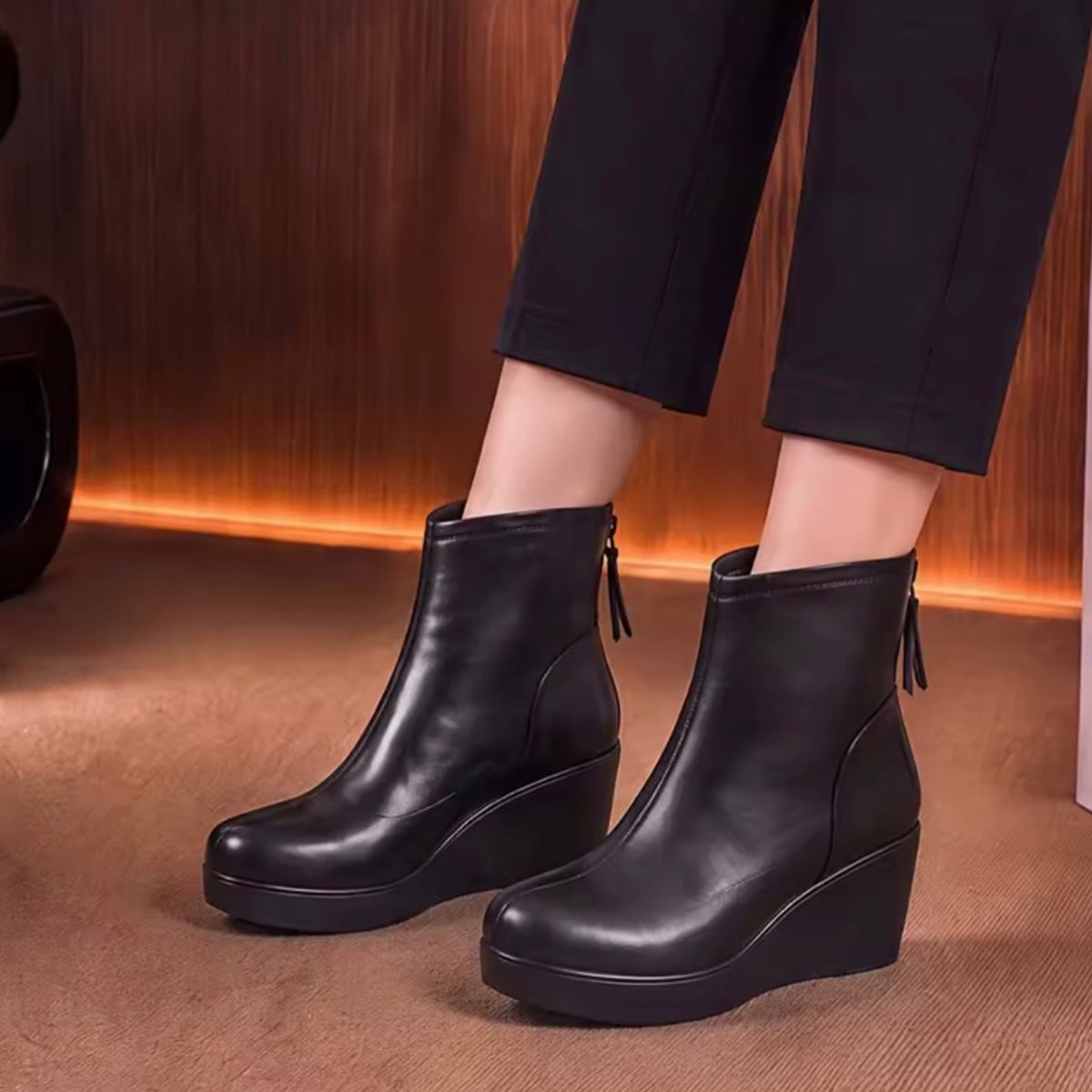 Black wedge ankle boots worn by a person standing on a brown floor against a dark background.
