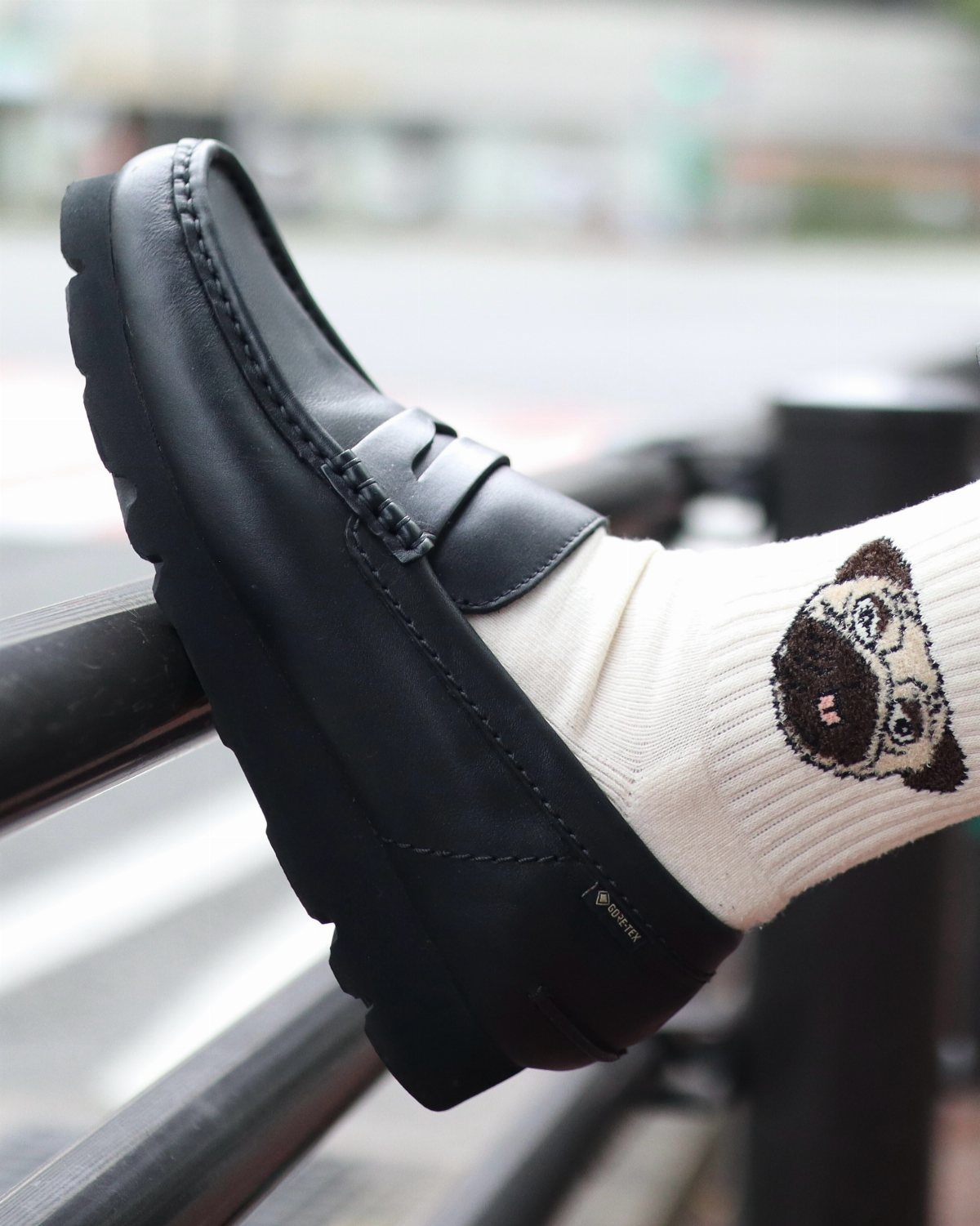 A black leather slip-on loafer worn with a pug patterned white sock on a metal railing.