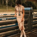A woman wearing a beige knit dress and patterned cowboy boots leaning against a metal gate.