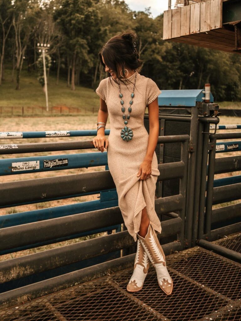 A woman wearing a beige knit dress and patterned cowboy boots leaning against a metal gate.