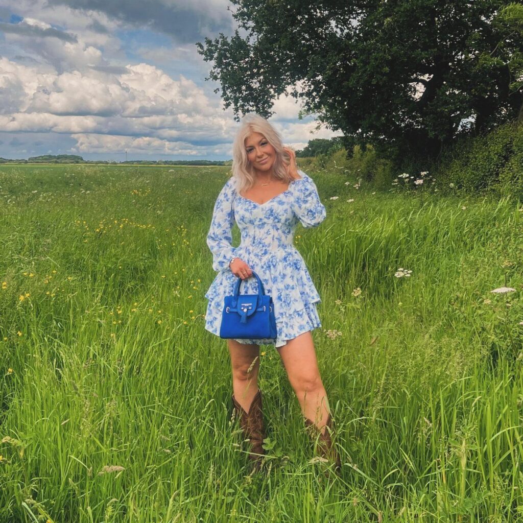 A blonde woman in a blue floral puff sleeve dress and brown western boots standing in a field.