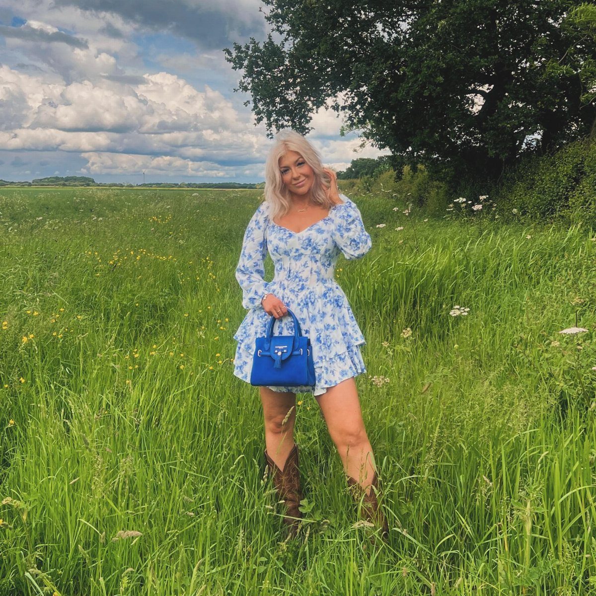 A blonde woman in a blue floral puff sleeve dress and brown western boots standing in a field.