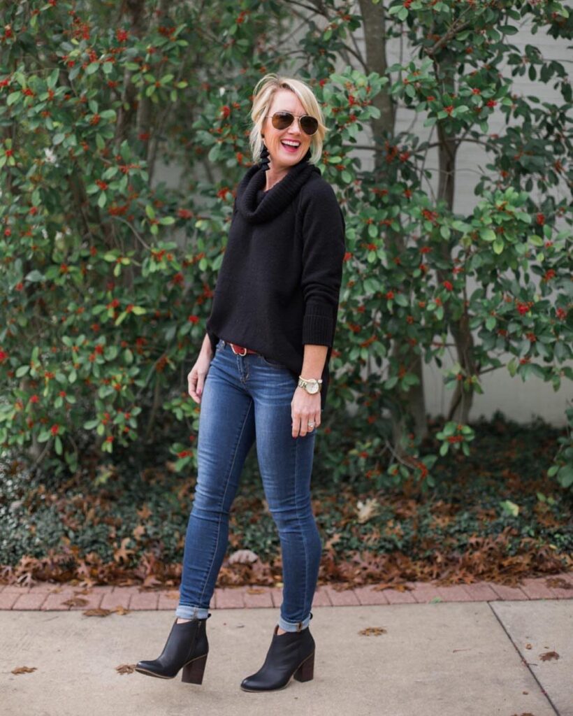 A woman wearing a black sweater, cuffed skinny jeans, and black heeled ankle boots standing outdoors.