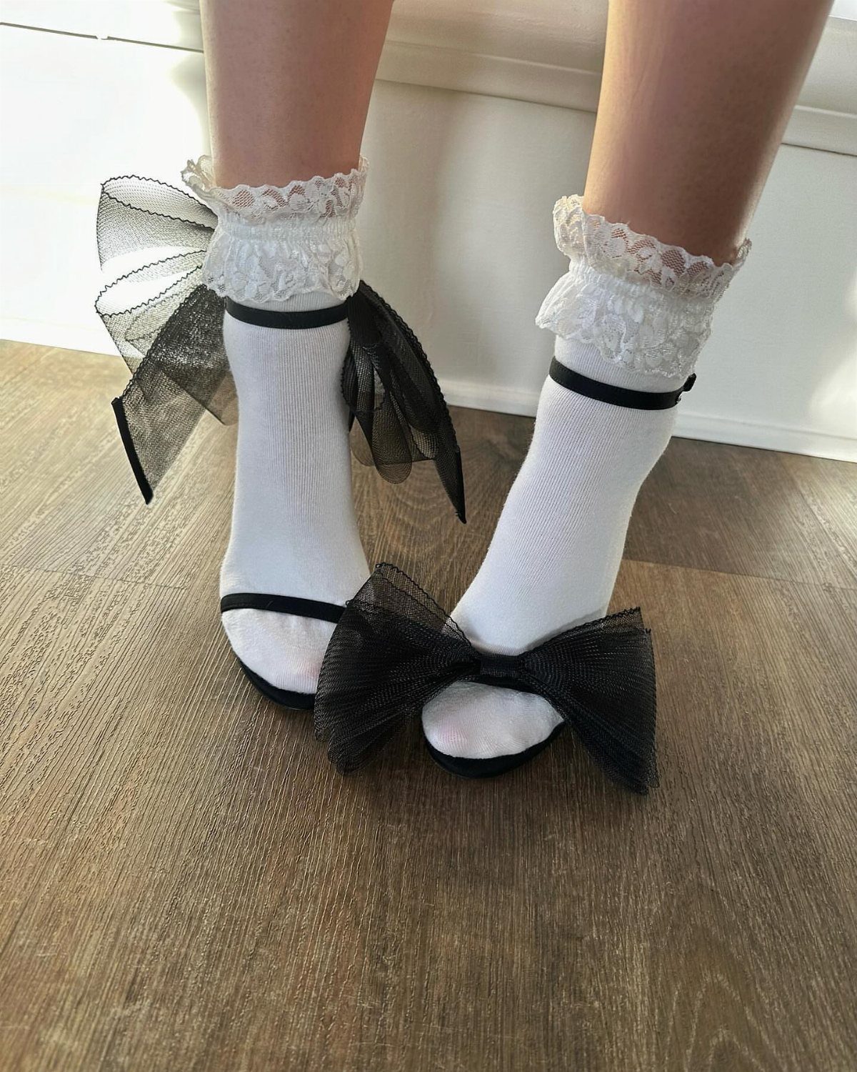 Feet wearing white lace socks and black high heels with large tulle bows on a wooden floor.