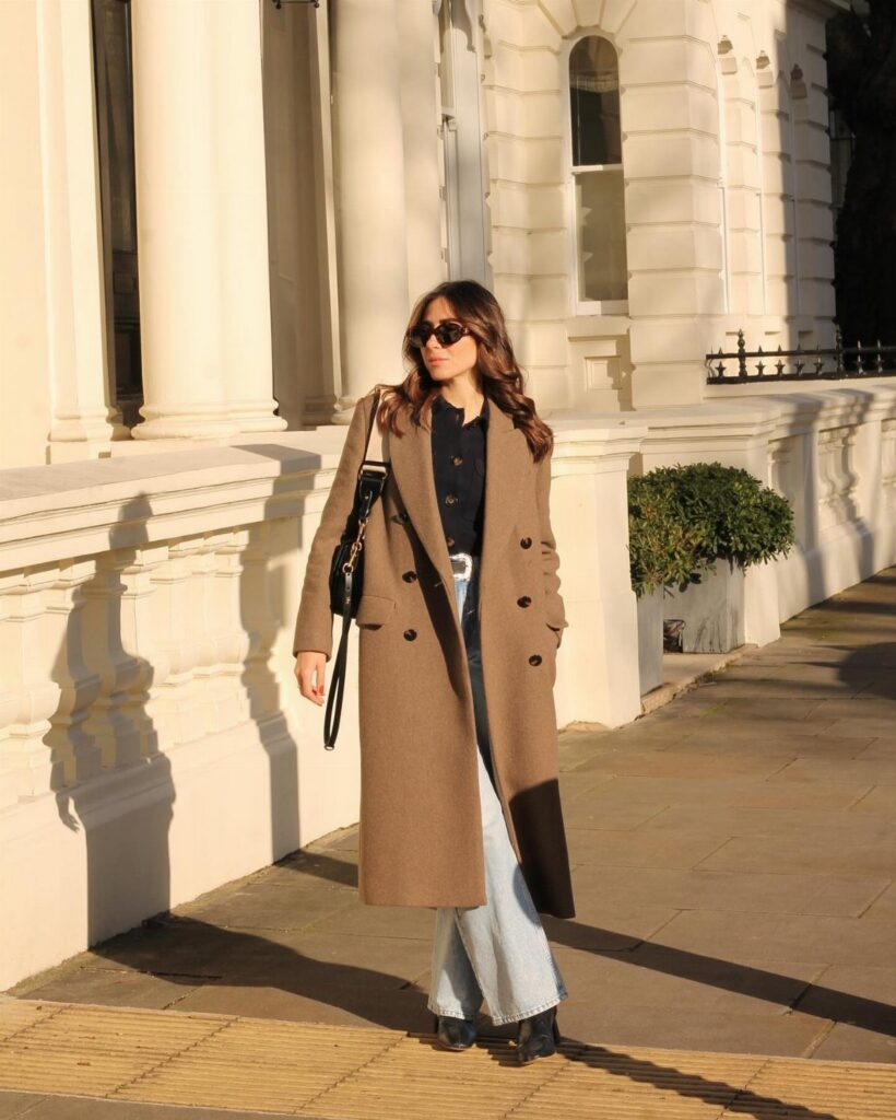 Woman in a long tan coat and wide-leg jeans wearing black ankle boots outdoors.