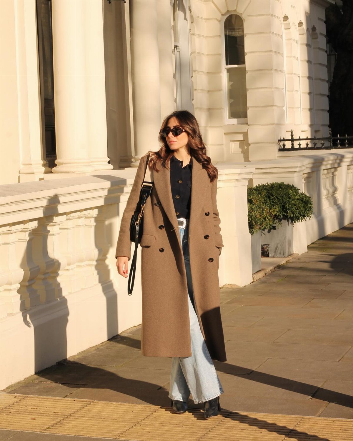 Woman in a long tan coat and wide-leg jeans wearing black ankle boots outdoors.