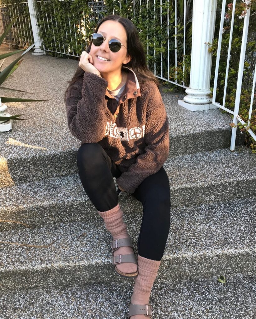 A woman sitting on steps wearing a brown fleece sweatshirt, black leggings, and Birkenstock sandals.
