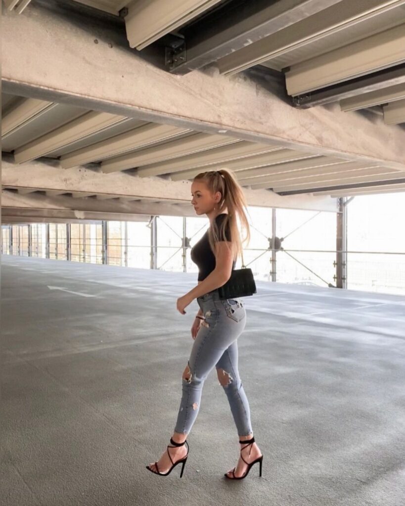A woman walking in a parking garage wearing ripped jeans and black high heels.