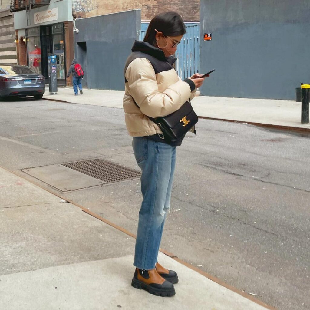 A woman in cropped jeans and Chelsea boots stands on a city sidewalk using her phone.