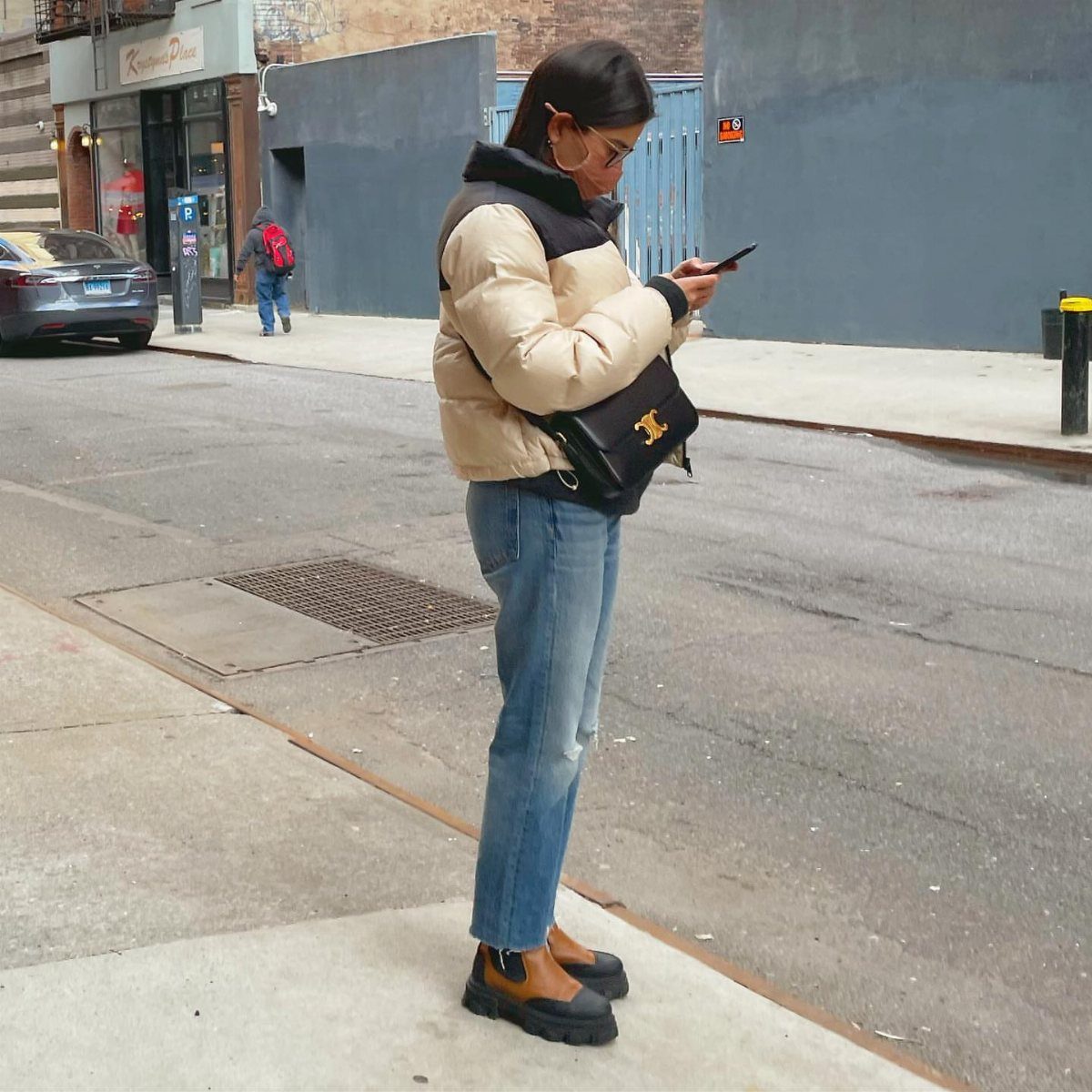 A woman in cropped jeans and Chelsea boots stands on a city sidewalk using her phone.
