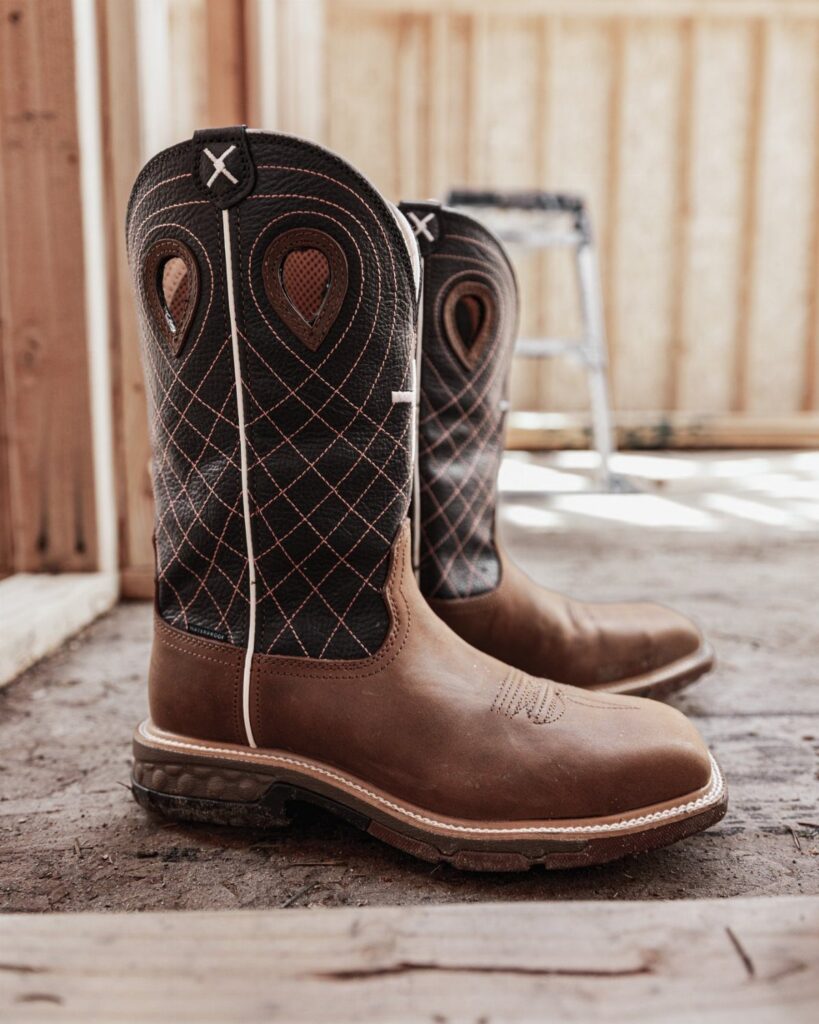 Brown leather cowboy boots with black patterned shafts sitting on a rustic wooden floor.