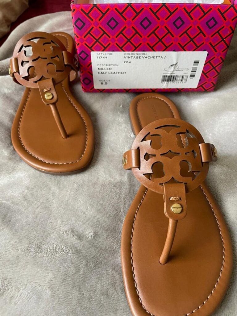 Tan leather Tory Burch Miller sandals resting next to their patterned shoebox on a grey surface.