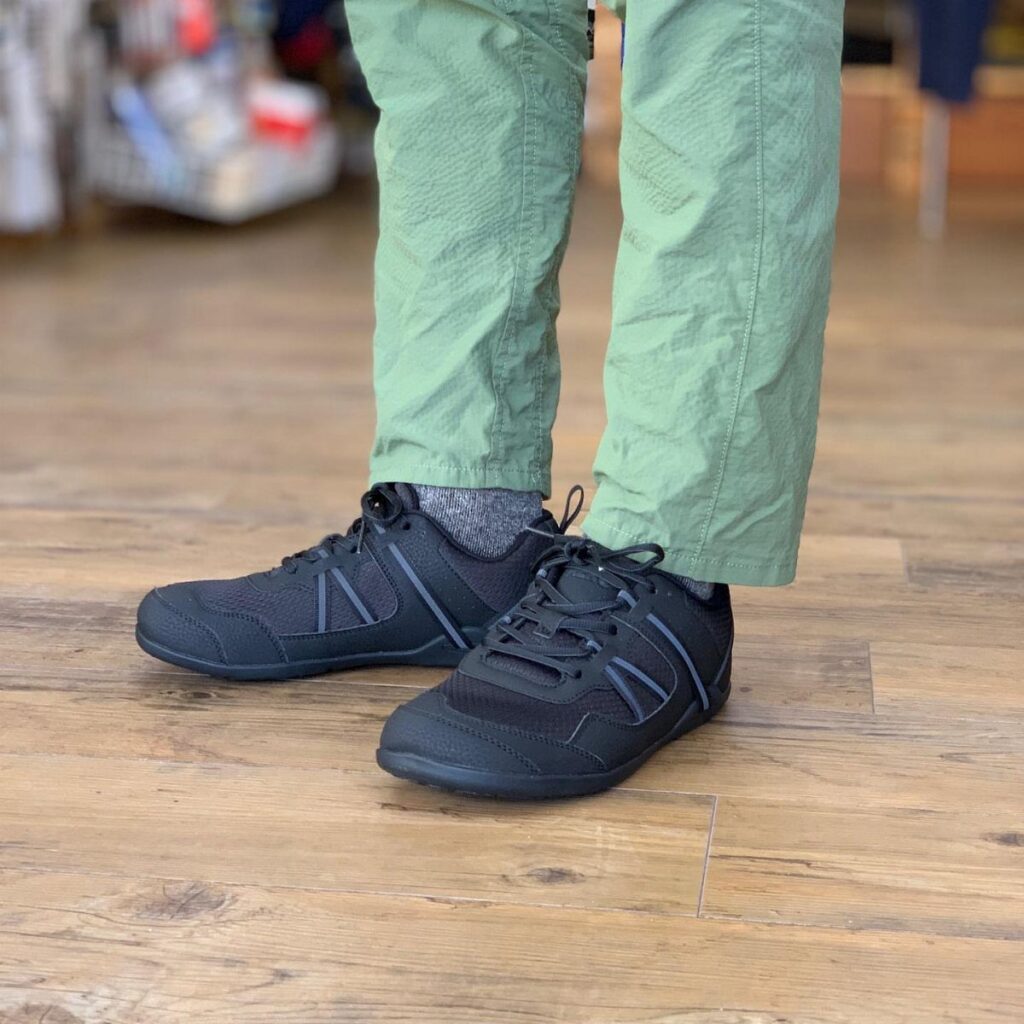 Black Xero Shoes Prio Original sneakers worn by a person in green trousers on a wooden floor.