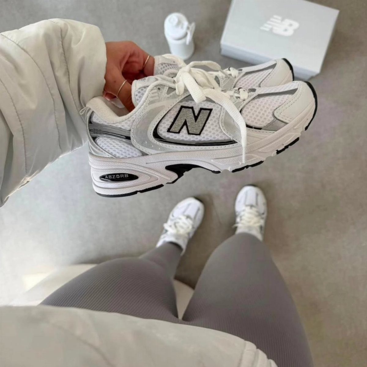 White and silver New Balance 530 sneakers held by a person wearing grey leggings.