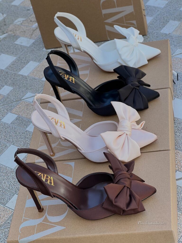Four pairs of pointed-toe stiletto sandals with large satin bows on cardboard boxes.