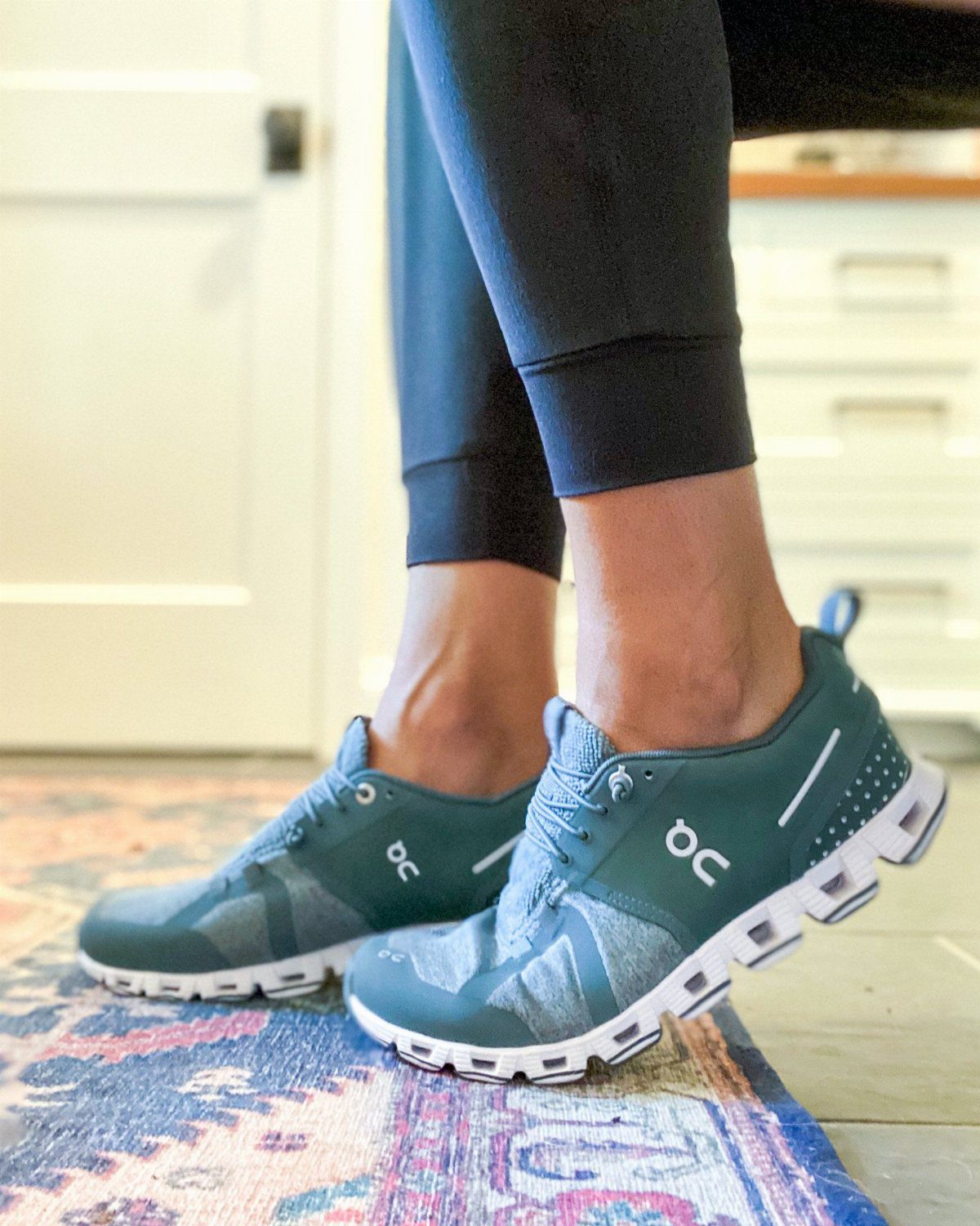 Teal On Cloud running shoes worn by a person sitting on a patterned rug.