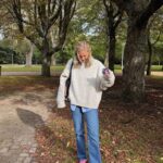 A woman wearing a cream oversized sweater, blue jeans, and pink sneakers standing in a park.