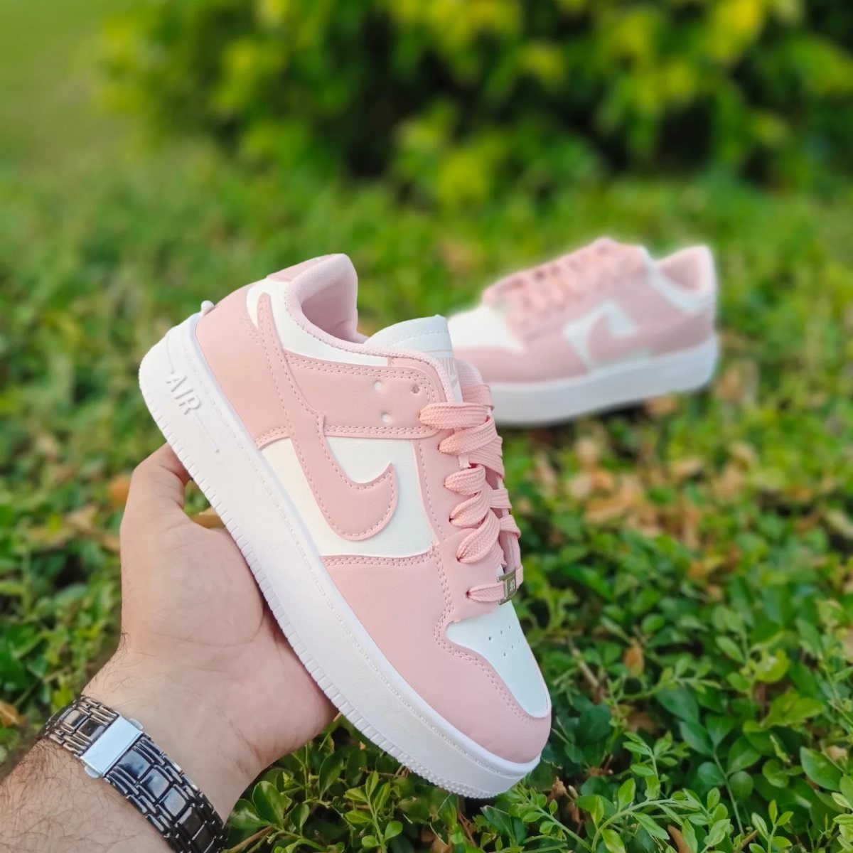 A hand holding a pink and white sneaker over a green grassy field.