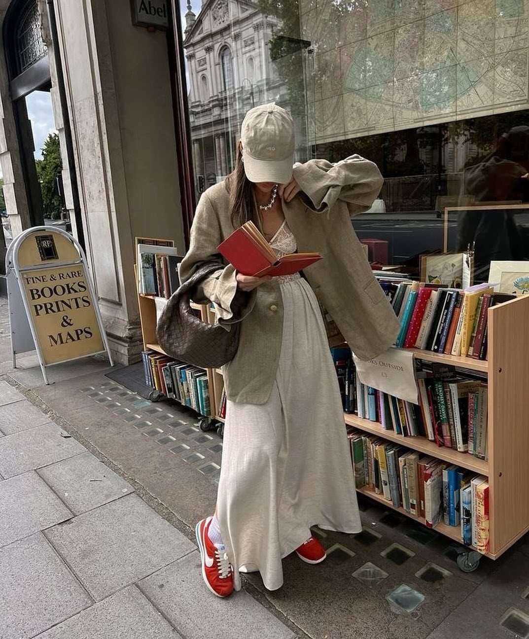 Woman in a long cream dress and red sneakers holding a red book outdoors.