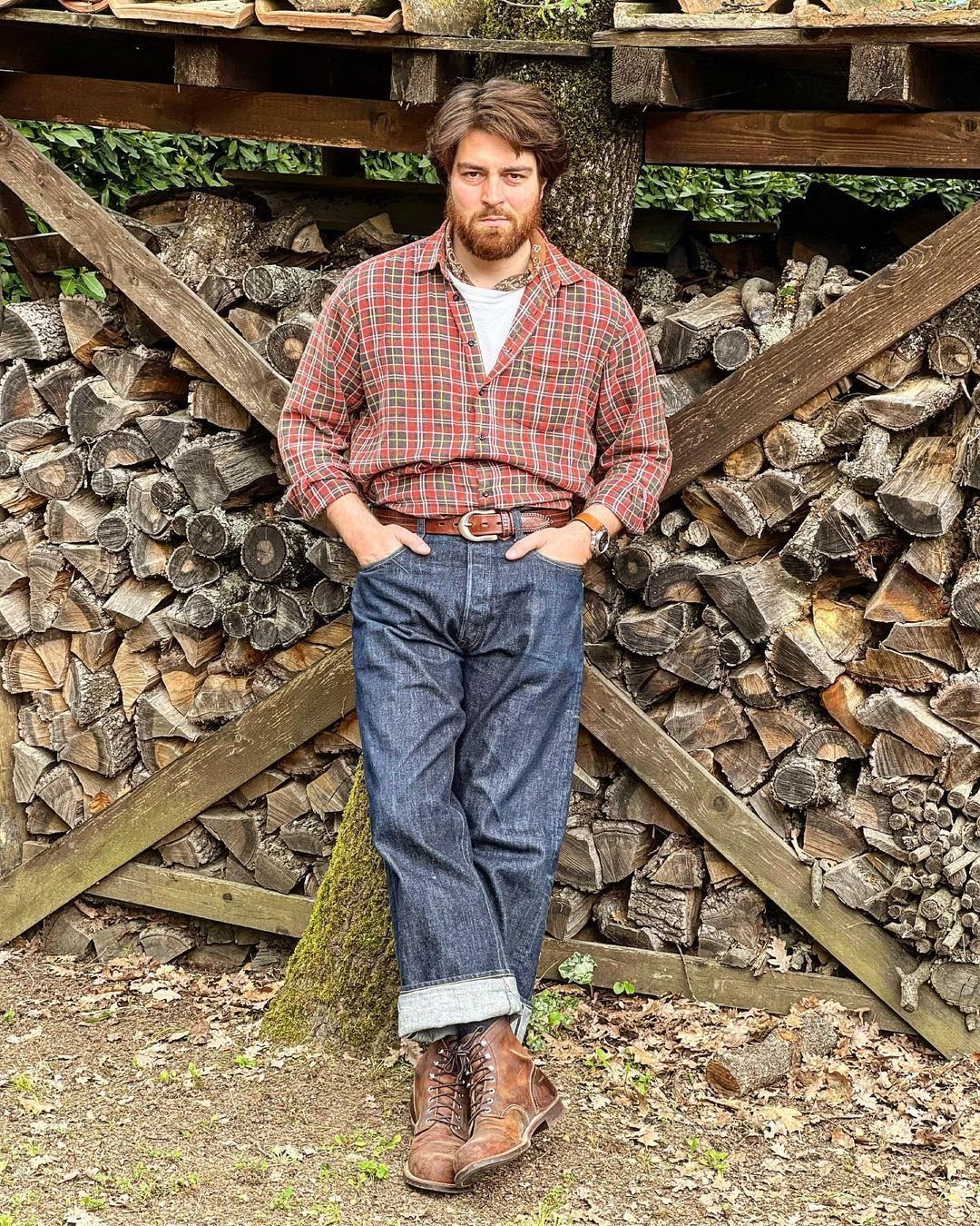 Man wearing a red plaid flannel shirt and dark denim jeans standing by a woodpile.