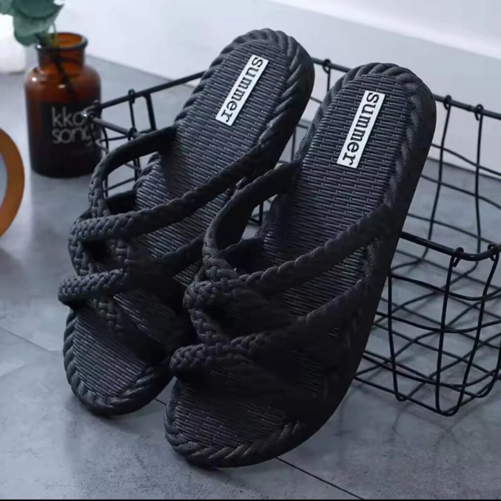 Black braided slide sandals with textured soles resting inside a black wire basket on a grey floor.