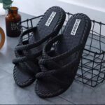 Black braided slide sandals with textured soles resting inside a black wire basket on a grey floor.