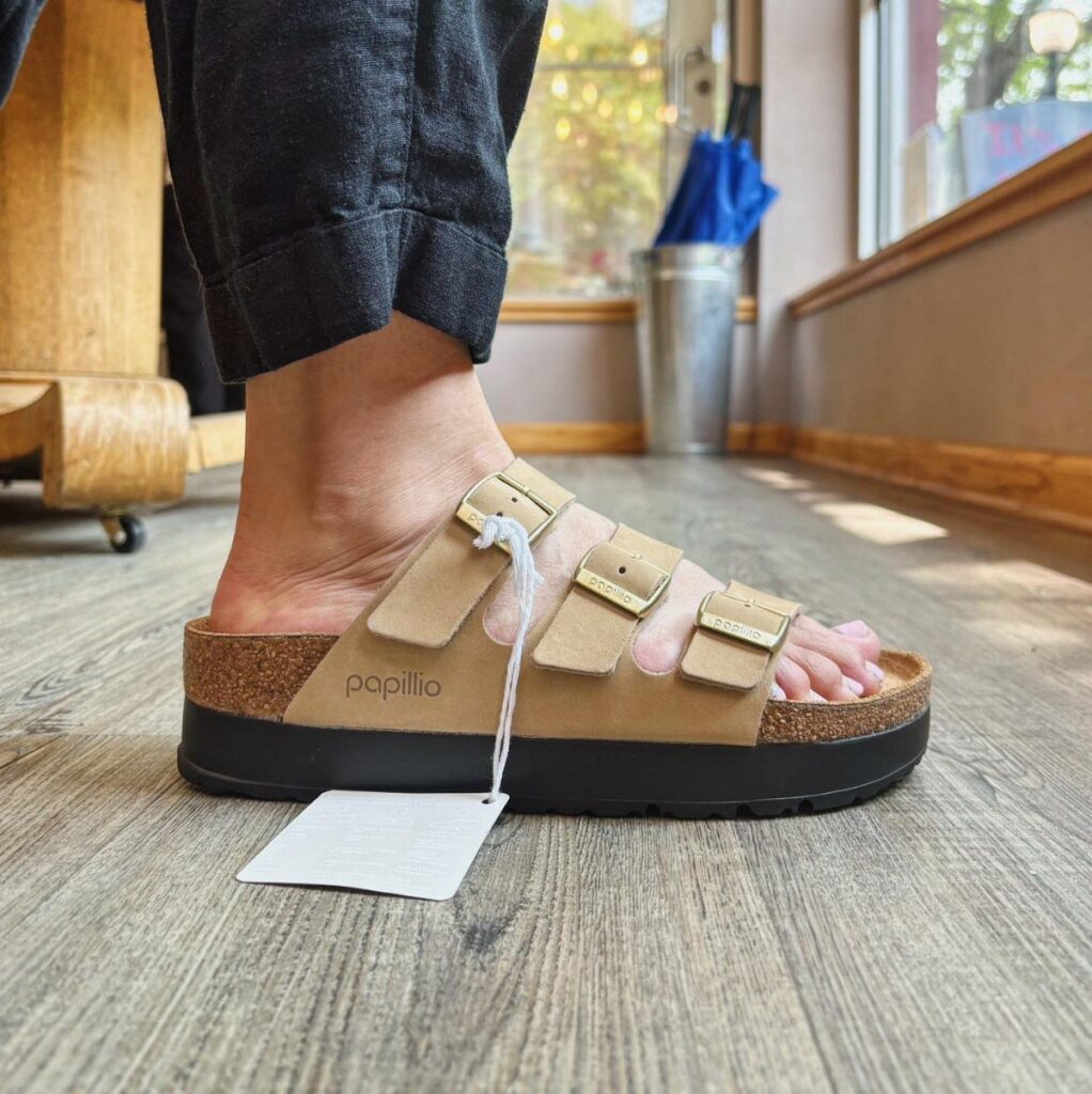 A person wearing tan two-strap sandals with a cork footbed and black sole on a wooden floor.