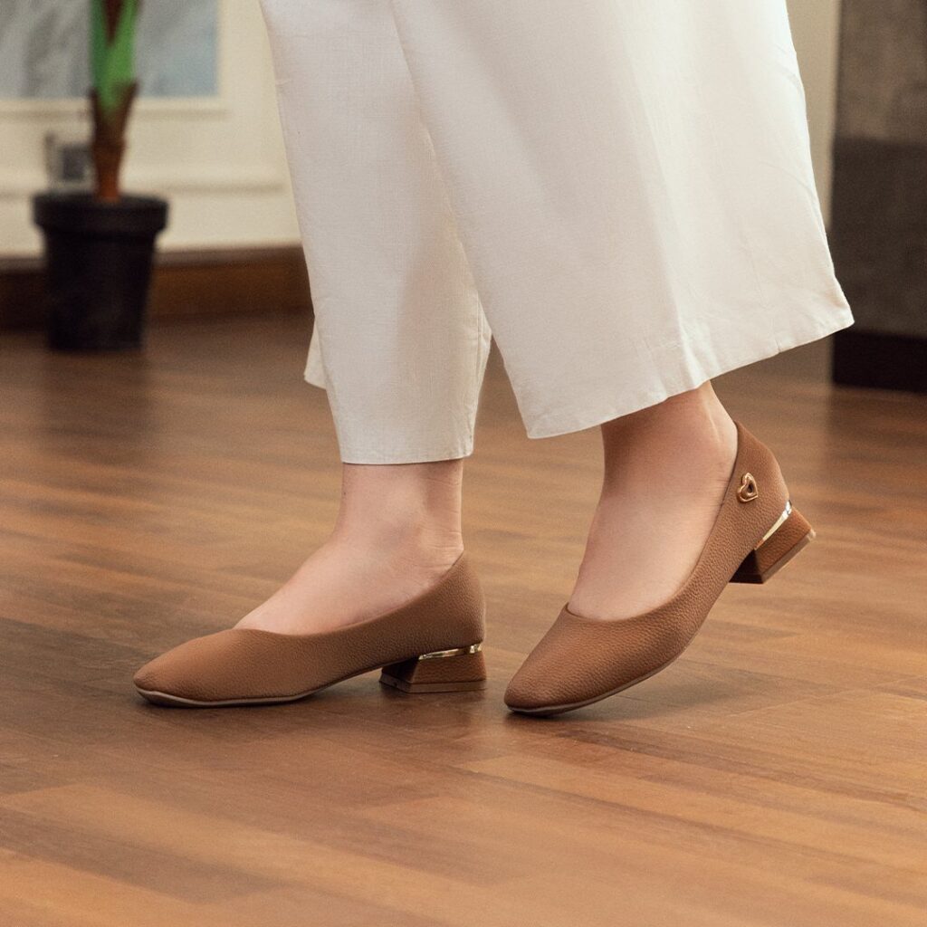 Brown pointed-toe flat shoes with metallic block heels worn by a person in white trousers.