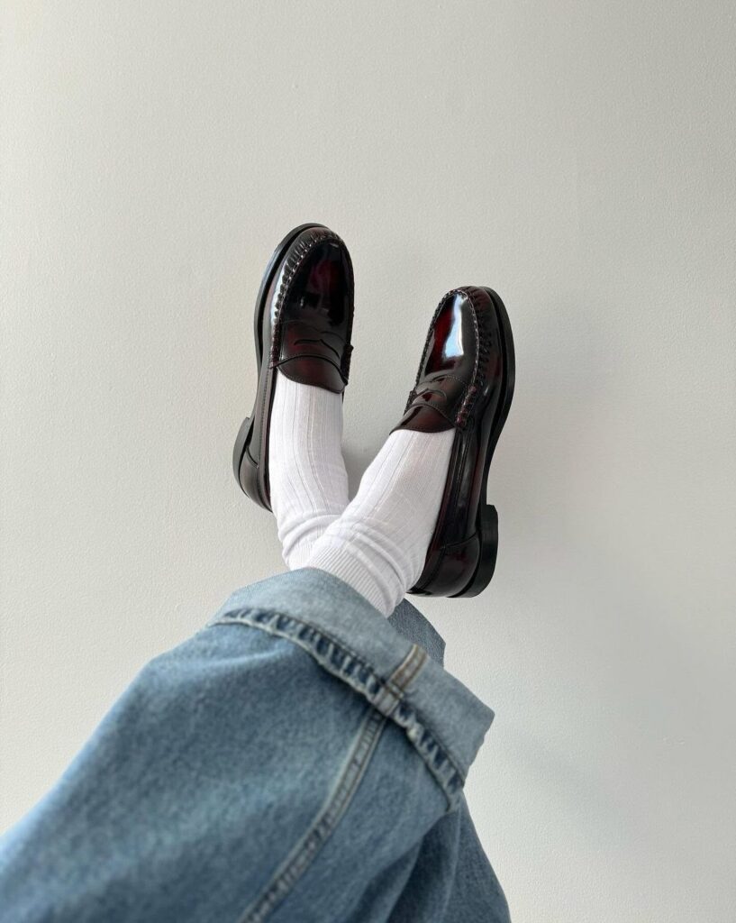 Dark burgundy patent leather loafers worn with white socks and blue denim jeans.