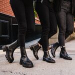 Three people walking on a sidewalk wearing black skinny jeans and various black leather boots.