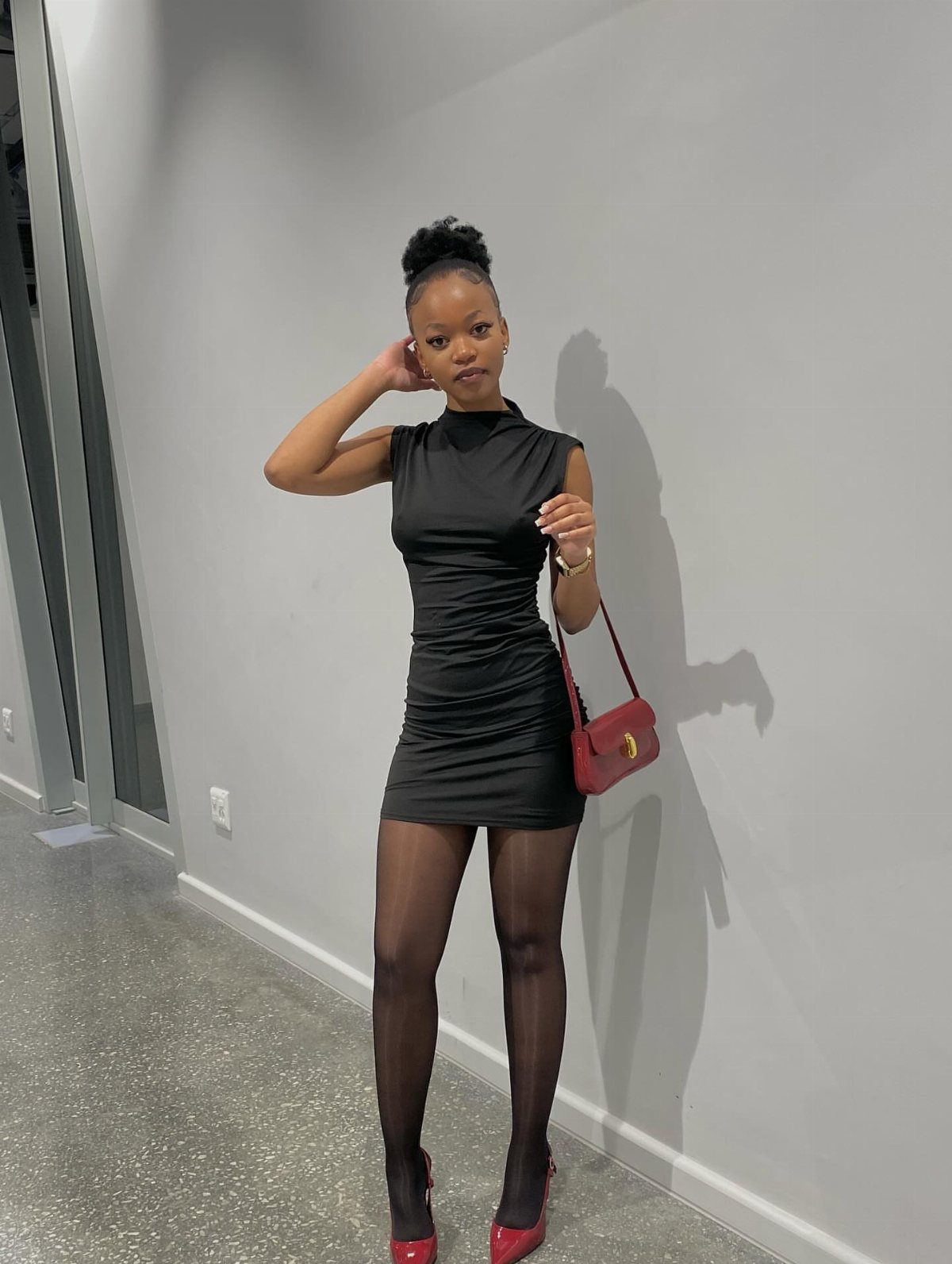 Woman in a black bodycon dress, black tights, and red pointed pumps posing against a wall.
