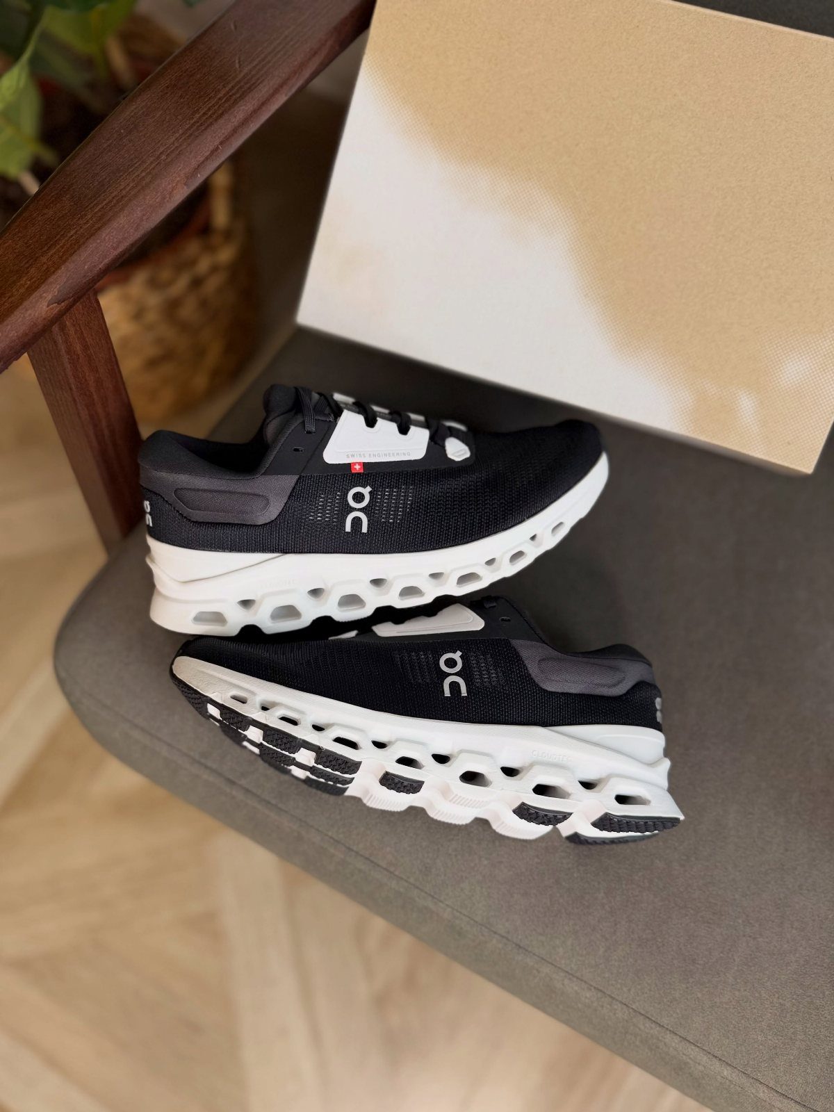 Black and white On Cloudstratus 3 running shoes sitting on a grey chair near a box.