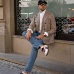 A man wearing a checkered blazer, blue jeans, and white sneakers sitting on a stone ledge.