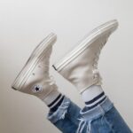 White high-top canvas sneakers worn with blue denim jeans and striped socks against a white background.
