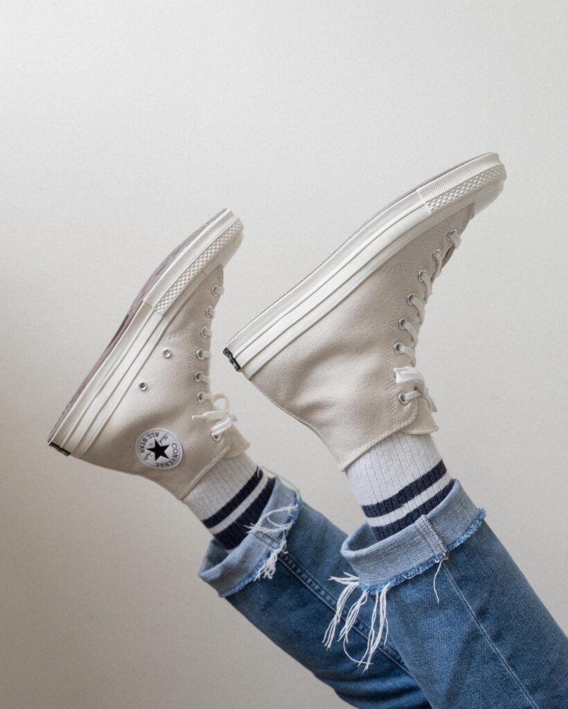 White high-top canvas sneakers worn with blue denim jeans and striped socks against a white background.