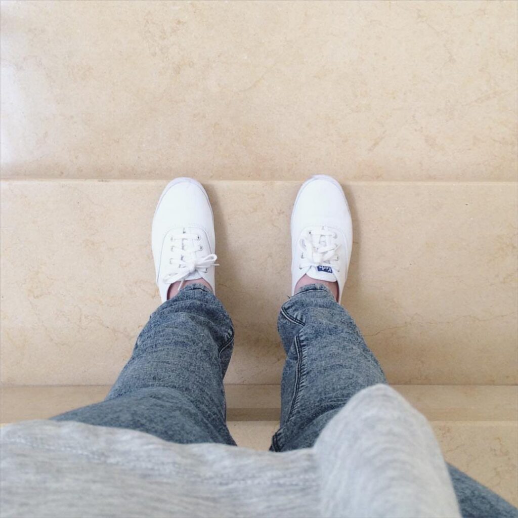 White sneakers paired with blue denim jeans and a grey sweater on beige stairs.