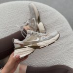 A person holding a beige Nike V2K Run sneaker against a white textured background.