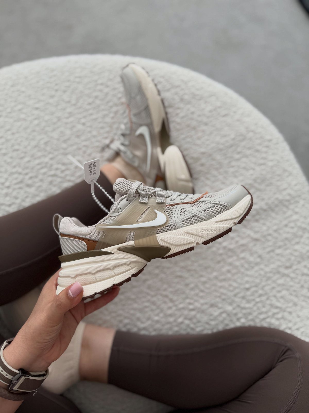 A person holding a beige Nike V2K Run sneaker against a white textured background.
