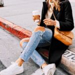Woman sitting on a curb wearing ripped light wash jeans and white leather sneakers.