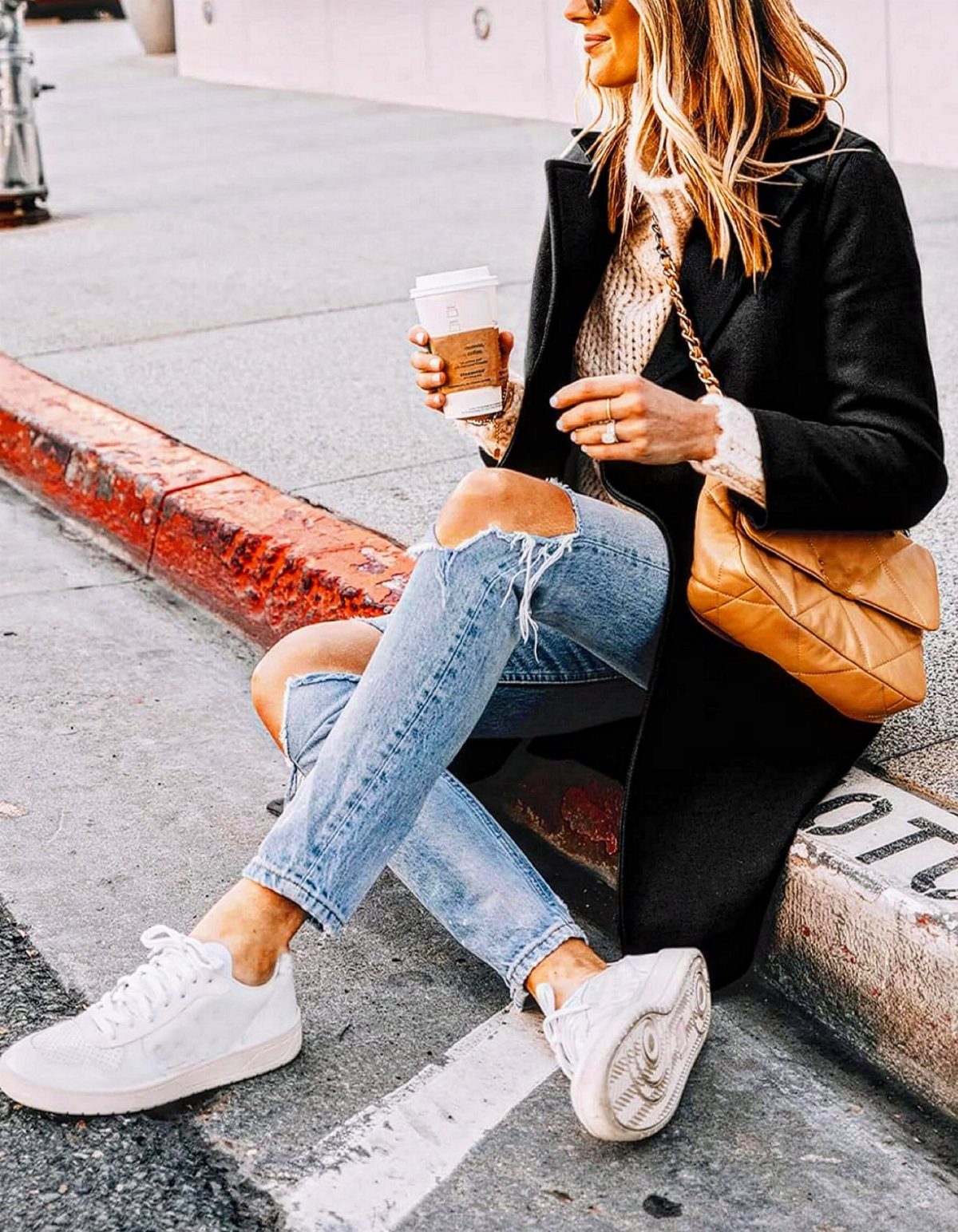 Woman sitting on a curb wearing ripped light wash jeans and white leather sneakers.