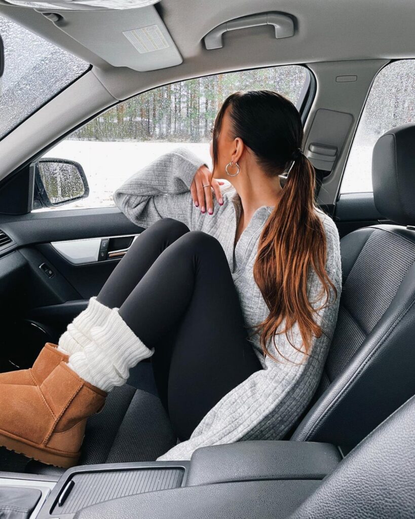 A woman wearing black leggings, white leg warmers, and tan Ugg boots sitting in a car.